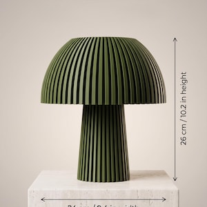 Puede incluir: Una l&aacute;mpara de mesa de color verde oliva oscuro con una pantalla en forma de seta y una base cil&iacute;ndrica, ambas con listones verticales. El texto DIMENSIONS aparece en la parte superior. La l&aacute;mpara mide 26 cm de alto y 24 cm de ancho.