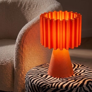 May include: A bright orange table lamp featuring a conical base and a cylindrical, vertically pleated lampshade. The lamp emits a warm, glowing light, highlighting the textured, rippled surface of the shade and the solid, smooth structure of the base.