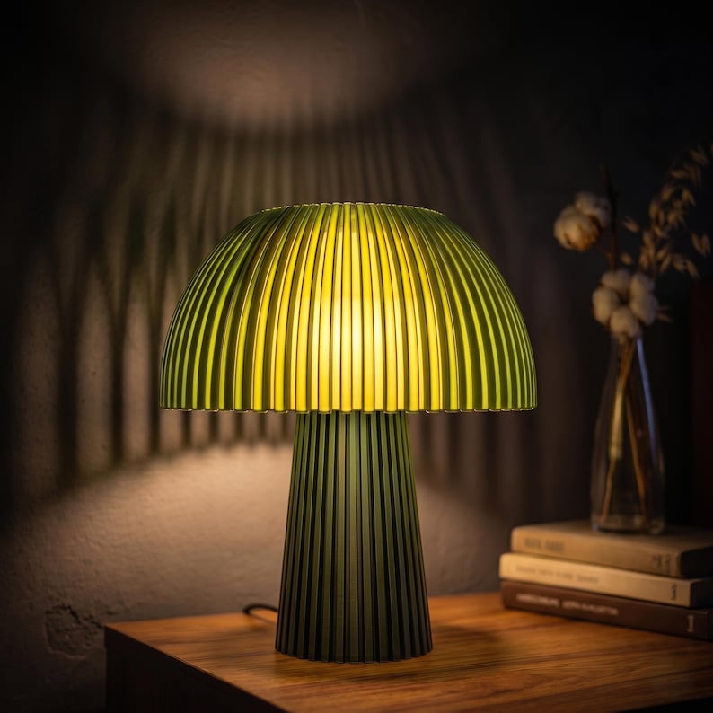Puede incluir: Una l&aacute;mpara de mesa con una pantalla acanalada en forma de hongo de color verde oliva y una base cil&iacute;ndrica a juego. La l&aacute;mpara emite un brillo amarillo c&aacute;lido y suave desde debajo de la pantalla, proyectando sombras verticales en la pared.