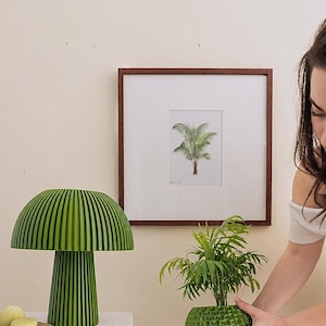 Puede incluir: Una l&aacute;mpara de mesa verde con forma de seta, dise&ntilde;o acanalado y una maceta verde texturizada que contiene una peque&ntilde;a palmera. La l&aacute;mpara presenta una pantalla ancha y redondeada con base cil&iacute;ndrica, mientras que la maceta tiene una superficie esf&eacute;rica con hoyuelos.