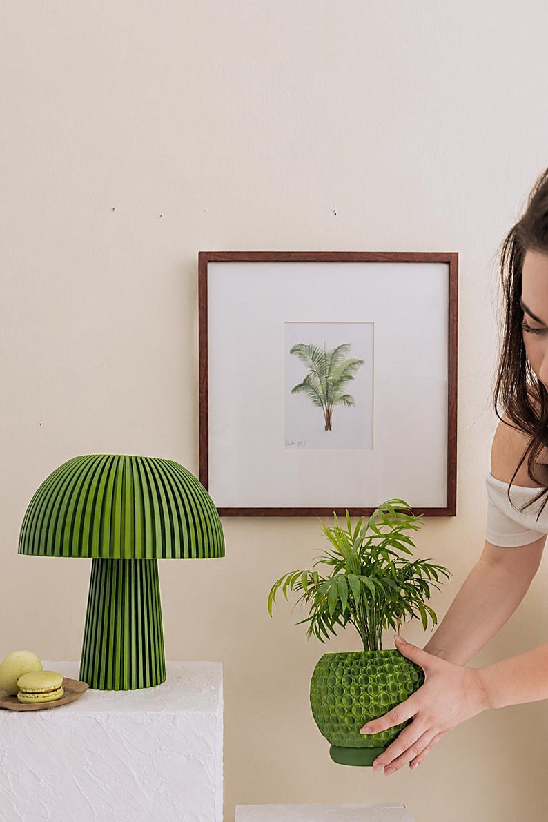 Puede incluir: Una l&aacute;mpara de mesa verde con forma de seta, dise&ntilde;o acanalado y una maceta verde texturizada que contiene una peque&ntilde;a palmera. La l&aacute;mpara presenta una pantalla ancha y redondeada con base cil&iacute;ndrica, mientras que la maceta tiene una superficie esf&eacute;rica con hoyuelos.