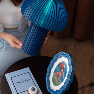 May include: A blue mushroom-shaped table lamp with a ribbed texture, a blue scalloped picture frame containing a photograph, a light blue book titled Little Book of Prada, and two small square and round candles with blue and white accents.