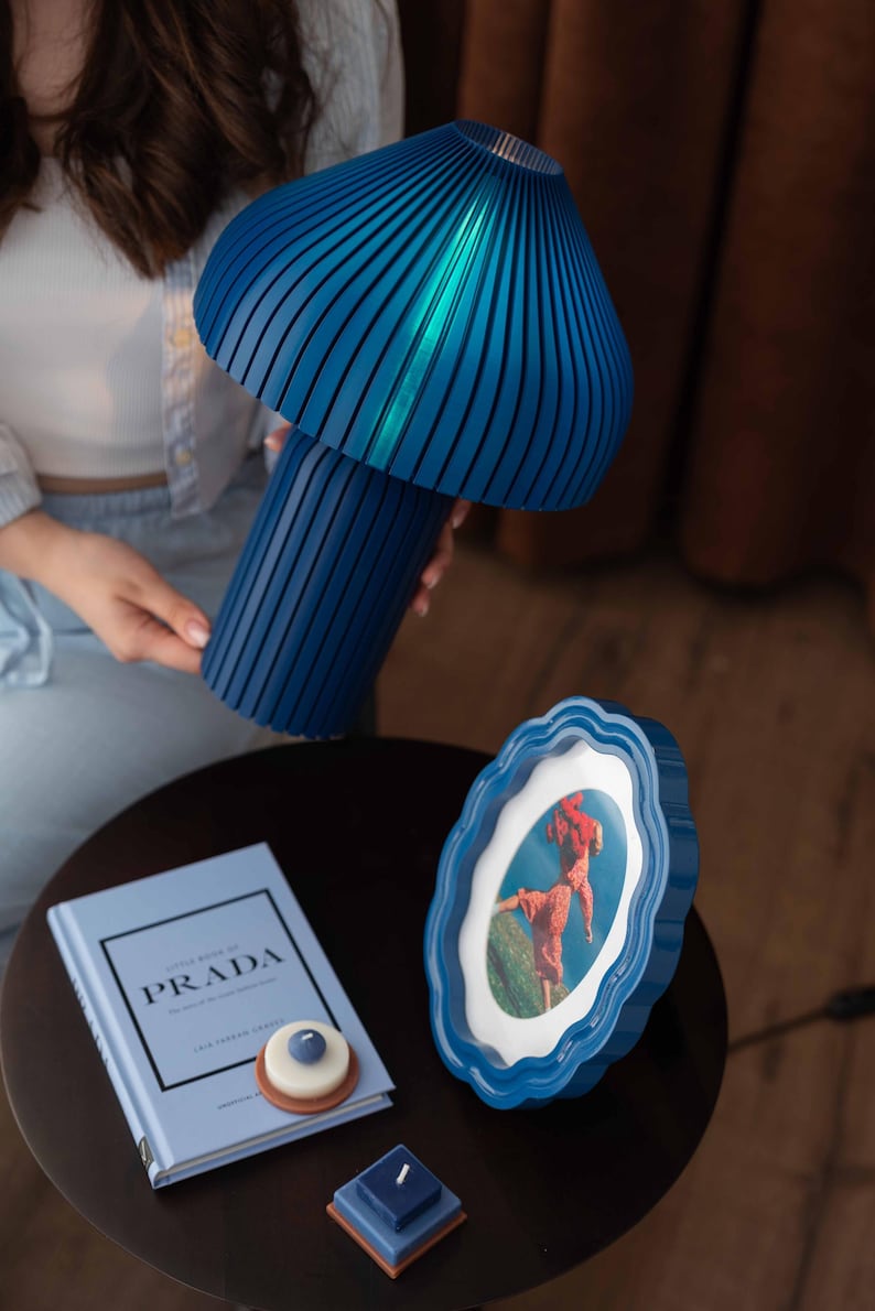 May include: A blue mushroom-shaped table lamp with a ribbed texture, a blue scalloped picture frame containing a photograph, a light blue book titled Little Book of Prada, and two small square and round candles with blue and white accents.