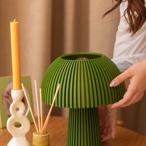 Puede incluir: Una l&aacute;mpara de mesa con forma de seta, con una pantalla y base de color verde oscuro y nervaduras verticales. Junto a ella hay una vela larga amarilla en un portavelas blanco abstracto y un difusor de varillas con palitos de madera.