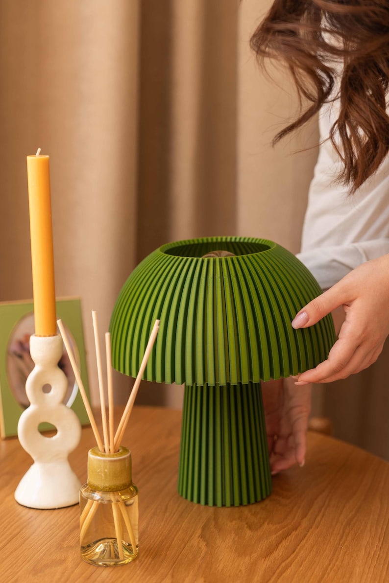Puede incluir: Una l&aacute;mpara de mesa con forma de seta, con una pantalla y base de color verde oscuro y nervaduras verticales. Junto a ella hay una vela larga amarilla en un portavelas blanco abstracto y un difusor de varillas con palitos de madera.