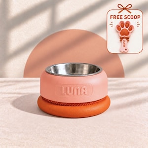 May include: A pink pet bowl with a stainless steel interior and the word LUNA embossed on the side, featuring a thick orange base. A small tag in the corner displays a paw-shaped scoop with the text FREE SCOOP and LUNA on the handle.