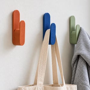 Puede incluir: Tres ganchos de pared en color naranja, azul y verde claro. Cada gancho presenta un diseño de doble punta con extremos redondeados. Un bolso de lona beige cuelga del gancho azul y una sudadera con capucha gris cuelga del gancho verde.
