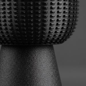 K&ouml;nnte beinhalten: Ein schwarzes zylindrisches Objekt mit einem texturierten, gitterartigen Muster im oberen Bereich und einem glatten, matten, konischen Sockel. Die Oberfl&auml;che weist ein sich wiederholendes geometrisches Design aus kleinen, erhabenen Rechtecken auf.