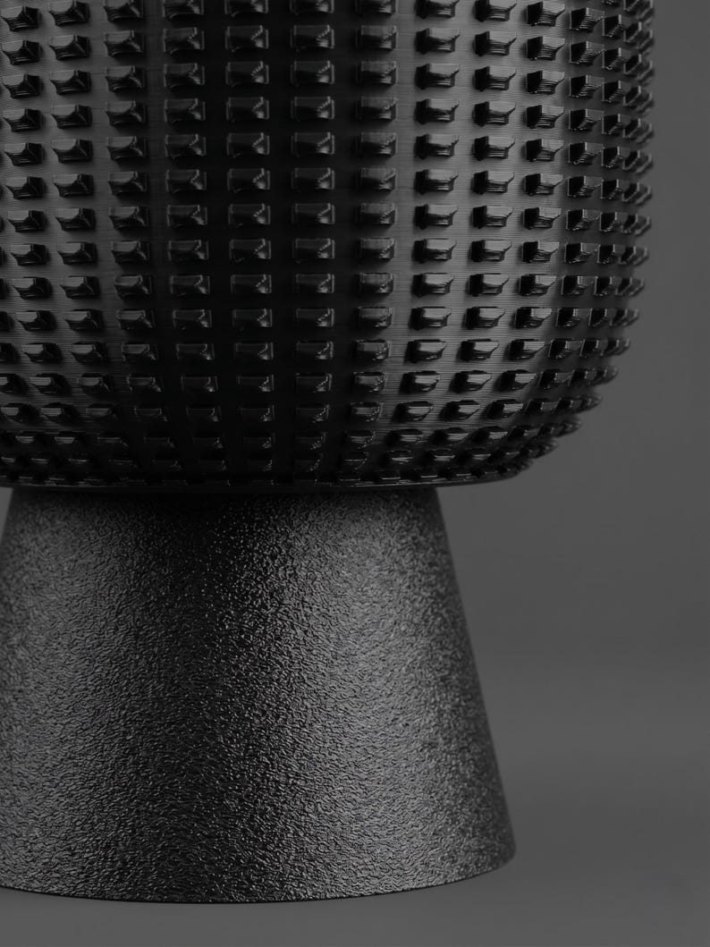 K&ouml;nnte beinhalten: Ein schwarzes zylindrisches Objekt mit einem texturierten, gitterartigen Muster im oberen Bereich und einem glatten, matten, konischen Sockel. Die Oberfl&auml;che weist ein sich wiederholendes geometrisches Design aus kleinen, erhabenen Rechtecken auf.