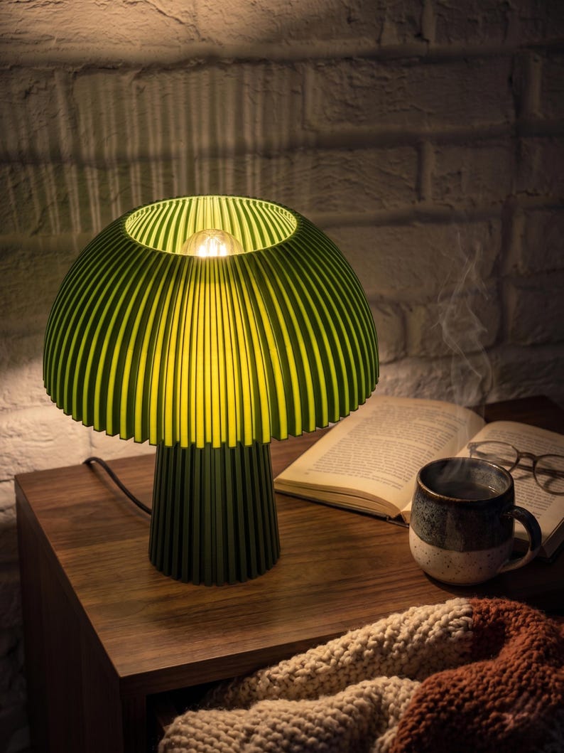 Puede incluir: Una l&aacute;mpara de mesa con forma de seta, con una pantalla acanalada de color verde lima y una base cil&iacute;ndrica de color verde oscuro. La l&aacute;mpara cuenta con una bombilla central que emite un brillo c&aacute;lido, iluminando el interior de la pantalla.