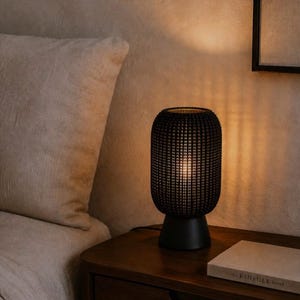 May include: A cylindrical table lamp with a black, textured, grid-patterned shade and a solid black base. The lamp emits a warm, soft glow from within, casting vertical light patterns onto the surrounding area. The design is simple and compact.