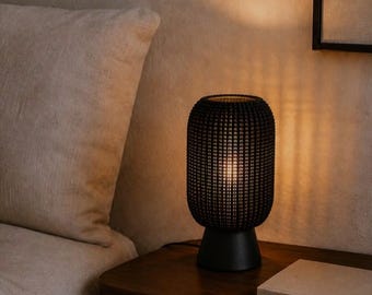 May include: A cylindrical table lamp with a black, textured, grid-patterned shade and a solid black base. The lamp emits a warm, soft glow from within, casting vertical light patterns onto the surrounding area. The design is simple and compact.