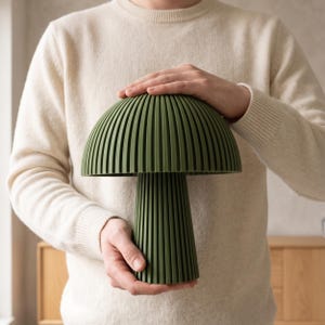 Puede incluir: Una l&aacute;mpara de mesa con forma de seta en un acabado verde oliva mate. La l&aacute;mpara presenta una c&uacute;pula ancha y una base cil&iacute;ndrica, ambas construidas con una serie de estr&iacute;as verticales paralelas que crean una apariencia texturizada y acanalada.