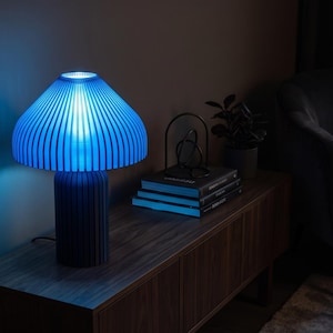 May include: A table lamp with a cylindrical base and a wide, mushroom-shaped shade, both featuring a vertical ribbed texture. The lamp emits a bright, vibrant blue light that illuminates the surrounding area with a distinct glow.