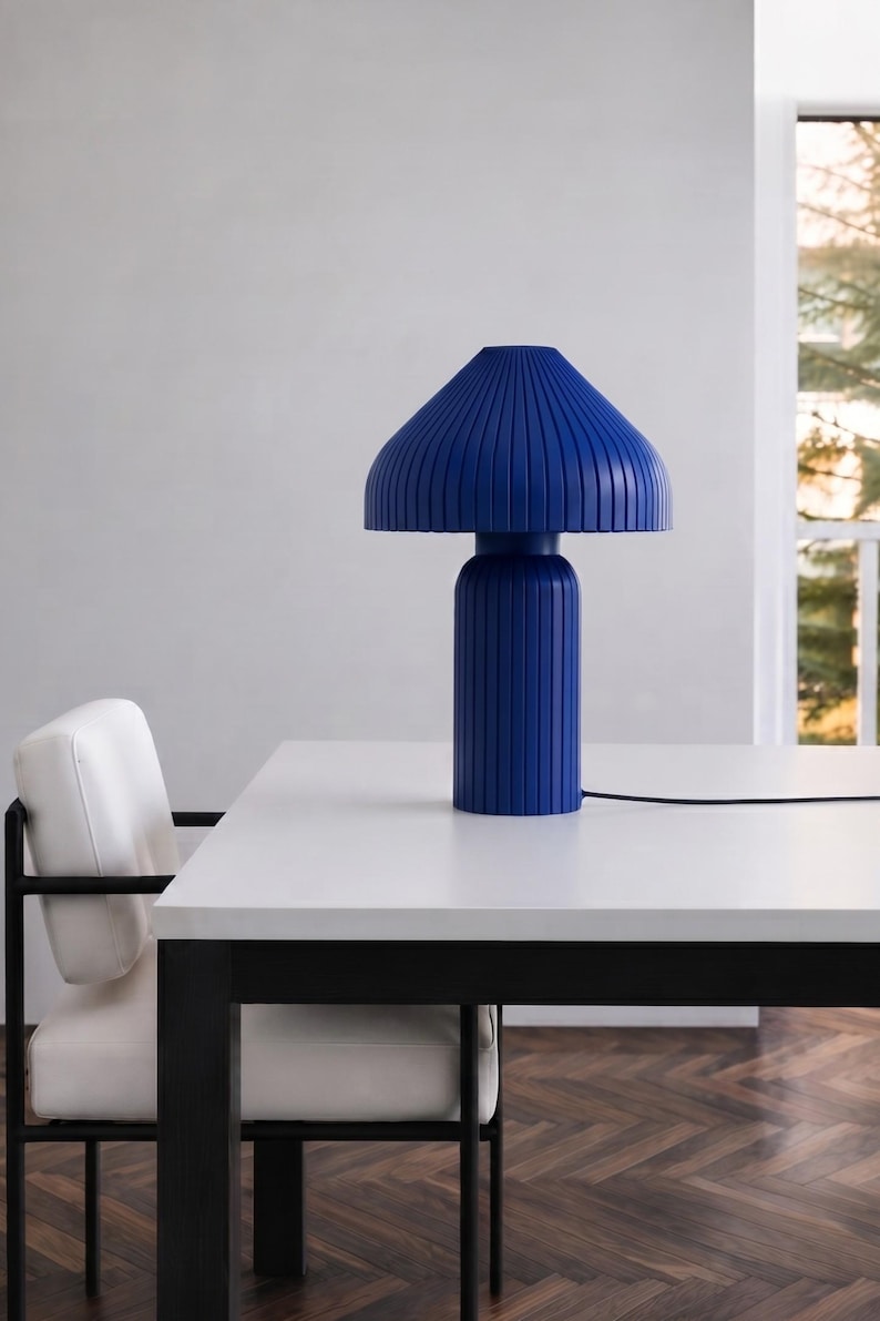 May include: A vibrant cobalt blue table lamp featuring a cylindrical base and a wide, conical shade, both characterized by a consistent vertical ribbed texture. The lamp includes a thin black power cord extending from the base to the side.