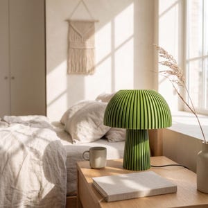 Puede incluir: Una l&aacute;mpara de mesa de color verde brillante con una pantalla en forma de seta y una textura acanalada vertical tanto en la pantalla como en la base cil&iacute;ndrica. La l&aacute;mpara tiene un cable de alimentaci&oacute;n negro y se apoya sobre una superficie de madera.