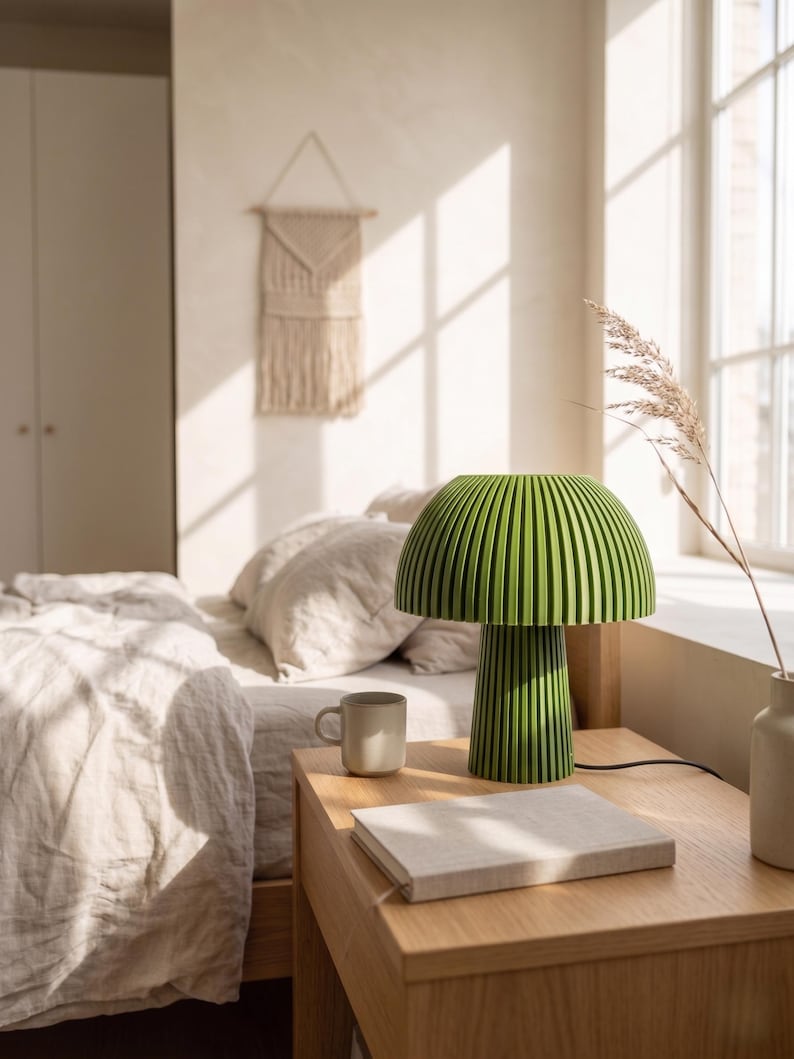 Puede incluir: Una l&aacute;mpara de mesa de color verde brillante con una pantalla en forma de seta y una textura acanalada vertical tanto en la pantalla como en la base cil&iacute;ndrica. La l&aacute;mpara tiene un cable de alimentaci&oacute;n negro y se apoya sobre una superficie de madera.