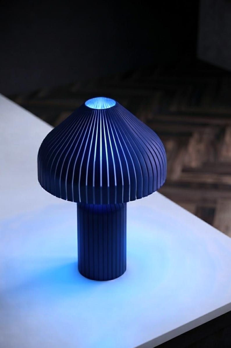May include: A dark blue table lamp with a mushroom-shaped shade and a cylindrical base, both featuring vertical slats. The lamp emits a bright blue light from the top opening and the base, casting a blue glow onto the surface below the lamp.