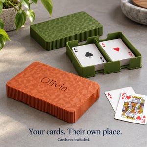 May include: Two rectangular playing card cases with a geometric textured surface and ribbed sides. One case is olive green, and the other is orange with the name Olivia embossed on top. An open green case holds two decks of cards.