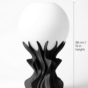 May include: A compact sculptural table lamp with a white globe shade and a black, stylized base. The lamp is 15 inches in height and 8.7 inches wide. The image includes the word "DIMENSIONS".