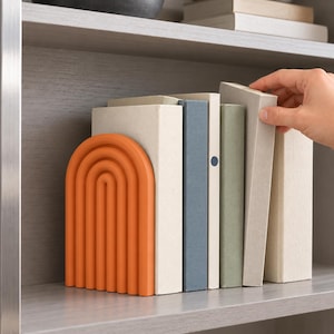 Puede incluir: Un sujetalibros naranja con forma de arco y diseño acanalado semicircular, colocado junto a una fila de libros con cubiertas de colores sólidos en tonos crema, azul y verde salvia, dispuestos verticalmente en un estante.