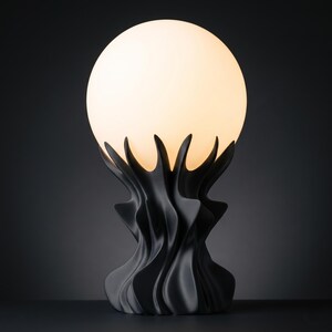 May include: A black table lamp with a unique design. The lamp features a spherical, off-white light bulb supported by a black, stylized base with a flowing, organic shape. The base has a series of upward-curving elements that cradle the bulb.