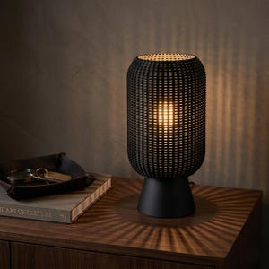 K&ouml;nnte beinhalten: Eine zylindrische Tischleuchte mit schwarzem, geflochtenem &Auml;u&szlig;eren und einem massiven, schwarzen, konischen Sockel. Die Lampe strahlt ein warmes Licht durch das Gittermuster und wirft deutliche, strahlenf&ouml;rmige Schatten auf die Fl&auml;che.