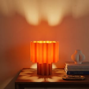以下が含まれることがあります： 垂直に溝が入った円筒形のシェードとベースを持つ、鮮やかなオレンジ色のテーブルランプ。ランプは温かみのある光を放ち、背後の壁に星形の影を落とすことで、部屋の中に際立った視覚的効果を生み出しています。