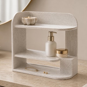 May include: A light grey speckled countertop organiser with two tiers and a curved side panel featuring vertical ribbed detailing. The top shelf holds a small flower-shaped candle, while the middle shelf displays a white pump bottle and a small jar.