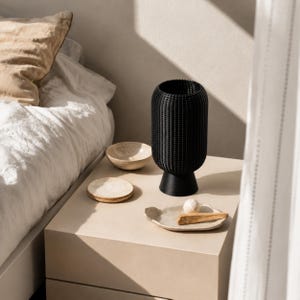 K&ouml;nnte beinhalten: Eine schwarze zylindrische Vase mit einer texturierten, gitterartigen Oberfl&auml;che, die auf einem ausgestellten Sockel steht. Die Vase wird von drei kleinen, hellen, unregelm&auml;&szlig;igen Keramikschalen begleitet, die eine kleine wei&szlig;e Kugel und einen Holzstab enthalten.