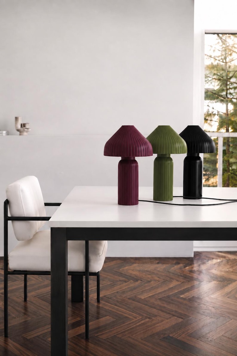 May include: Three table lamps with a mushroom-shaped silhouette and a vertical ribbed texture. The lamps are colored in deep burgundy, olive green, and black, each featuring a cylindrical base and a wide, flared shade with a black power cord.