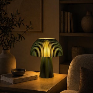 Puede incluir: Una l&aacute;mpara de mesa de color verde oscuro con una pantalla en forma de seta y una base cil&iacute;ndrica, ambas con un dise&ntilde;o de listones verticales. La l&aacute;mpara emite un brillo amarillo c&aacute;lido y suave desde debajo de la pantalla, iluminando el &aacute;rea.