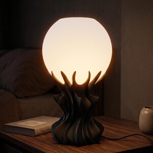 May include: A modern table lamp with a spherical, frosted glass shade. The lamp's base has a dark, organic design, resembling stylized flames or petals. The lamp is on a wooden nightstand next to a book and a small vase.