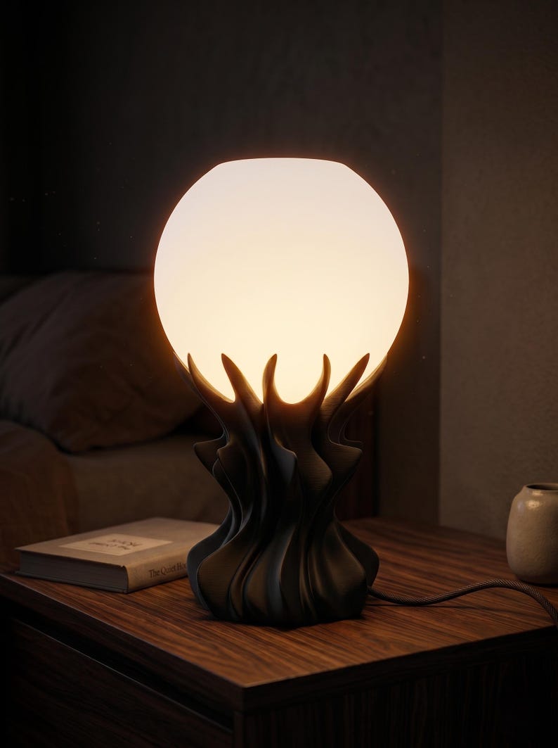 May include: A modern table lamp with a spherical, frosted glass shade. The lamp's base has a dark, organic design, resembling stylized flames or petals. The lamp is on a wooden nightstand next to a book and a small vase.