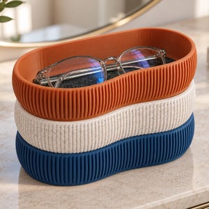 Pode incluir: Uma pilha de três tabuleiros de armazenamento ovais com paredes exteriores caneladas. O tabuleiro superior é cor de laranja queimado, o do meio é branco sujo salpicado e o inferior é azul-marinho. Óculos com armação transparente repousam no tabuleiro.