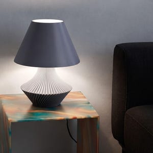 Könnte beinhalten: Eine Tischleuchte mit einem dunkelgrauen, konischen Lampenschirm und einem weißen, gerippten, ausgestellten Sockel. Die Lampe strahlt ein sanftes Licht unter dem Schirm aus, das die texturierte, gefaltete Oberfläche des Sockels beleuchtet.