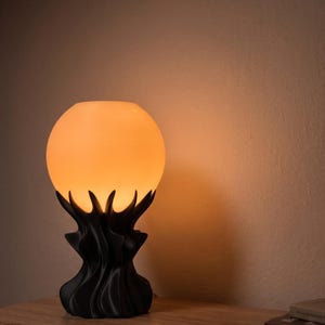 May include: A decorative table lamp with a spherical, glowing orange lampshade. The base is black and features a stylized, organic design. The lamp is placed on a wooden surface.
