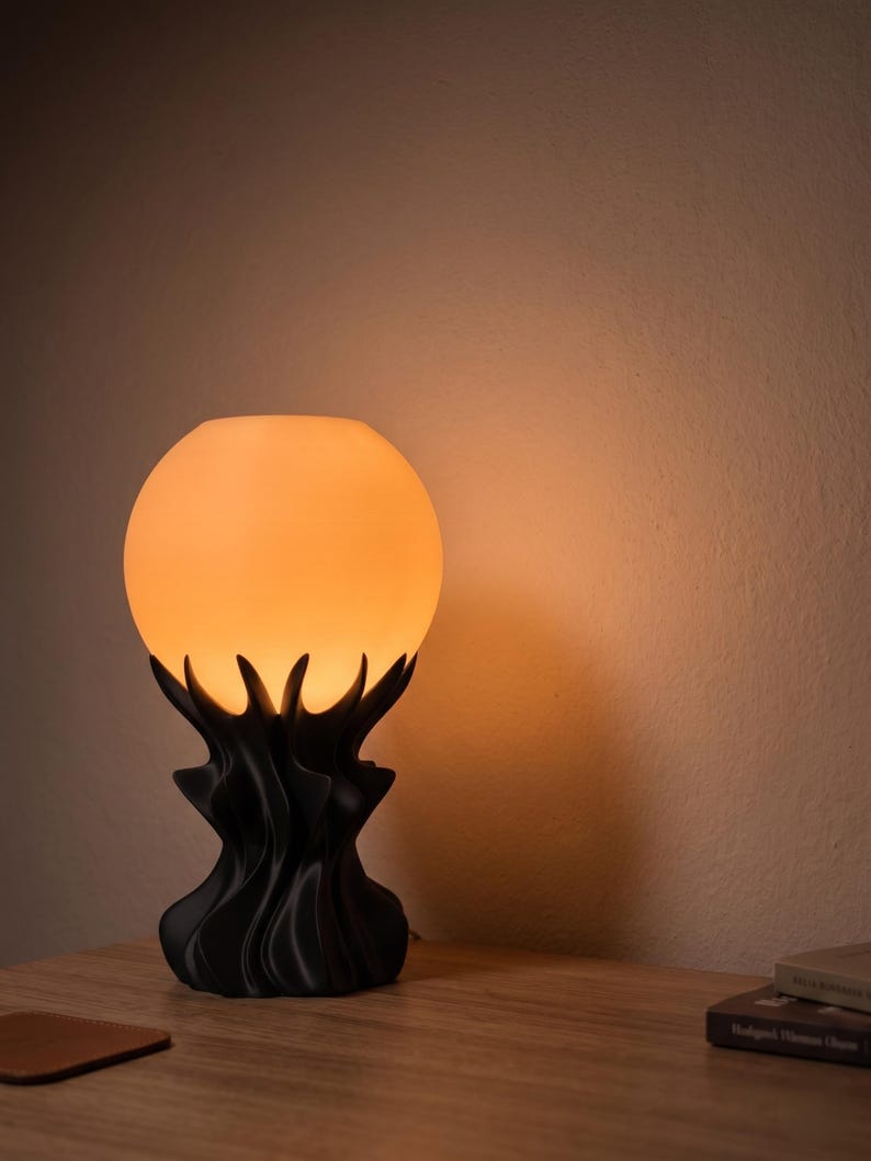 May include: A decorative table lamp with a spherical, glowing orange lampshade. The base is black and features a stylized, organic design. The lamp is placed on a wooden surface.