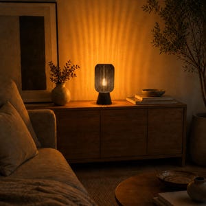 K&ouml;nnte beinhalten: Eine zylindrische Tischleuchte mit einem dunklen, lamellierten Au&szlig;enschirm, der ein warmes, leuchtendes Licht abgibt. Die Lampe verf&uuml;gt &uuml;ber einen massiven, abgerundeten Sockel und wirft vertikale Lichtmuster an die Wand, was f&uuml;r eine sanfte Beleuchtung sorgt.