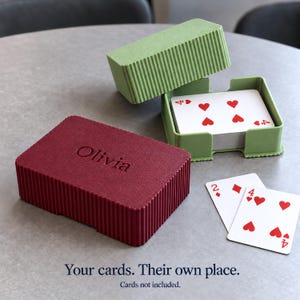 May include: A maroon rectangular card box with the name Olivia embossed on the lid, featuring a ribbed texture. A matching light green open card holder contains a deck of playing cards, with two additional cards placed nearby.