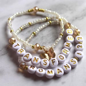 May include: Three beaded bracelets. One bracelet is made of small white beads and gold accents. The second bracelet is made of white letter beads that spell out the word "TRANSFORM". The third bracelet is made of white letter beads that spell out the word "2022". A gold butterfly charm hangs from the "TRANSFORM" bracelet.