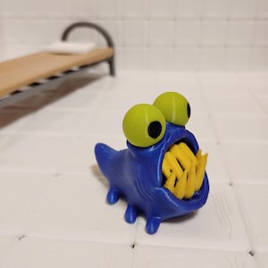 May include: A small blue creature figurine with a textured body, four short legs, and a wide open mouth revealing sharp yellow teeth. The creature has two large, round, light green eyes with black pupils positioned on top of its head.