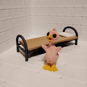 May include: A pink, cartoon-like duck figurine with large, round eyes and yellow feet stands in front of a miniature bed. The bed has a wooden platform, black frame, and a small white pillow. The background is a white tiled wall.