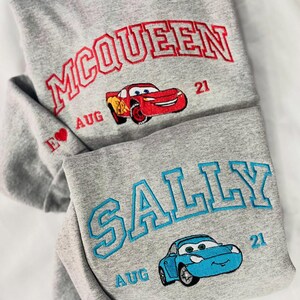 Pode incluir: Dois hoodies cinzentos com desenhos bordados. Um apresenta "MCQUEEN" em vermelho, um gr&aacute;fico de carro vermelho e "AUG 21". O outro tem "SALLY" em azul e um gr&aacute;fico de carro azul, com "AUG 21".