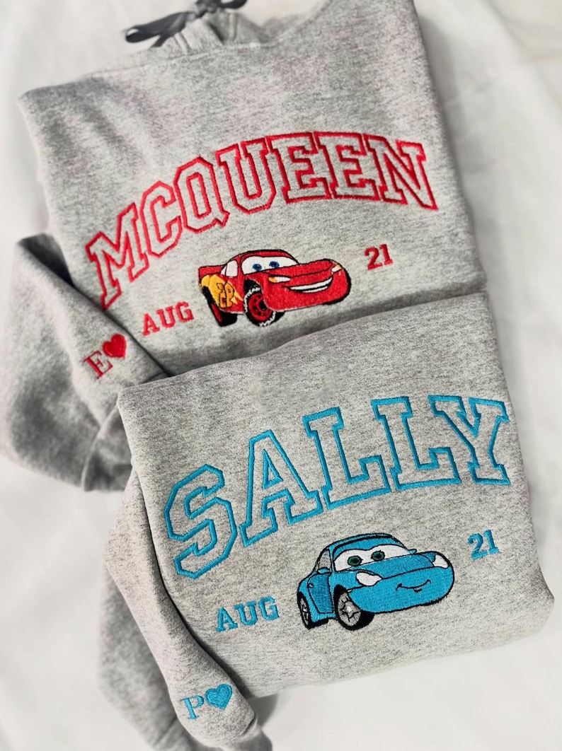 Pode incluir: Dois hoodies cinzentos com desenhos bordados. Um apresenta "MCQUEEN" em vermelho, um gr&aacute;fico de carro vermelho e "AUG 21". O outro tem "SALLY" em azul e um gr&aacute;fico de carro azul, com "AUG 21".