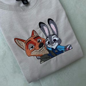 May include: A light beige t-shirt featuring an embroidered design of Nick Wilde, a fox, and Judy Hopps, a rabbit, from the animated film Zootopia. The characters are depicted in their signature outfits, with vibrant colors and detailed stitching.