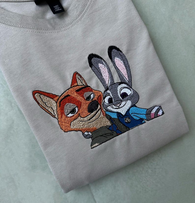 May include: A light beige t-shirt featuring an embroidered design of Nick Wilde, a fox, and Judy Hopps, a rabbit, from the animated film Zootopia. The characters are depicted in their signature outfits, with vibrant colors and detailed stitching.