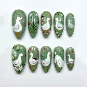 May include: A set of ten almond-shaped false nails. Each nail is painted with a light green base and features a white goose design. Some nails have additional details like flowers, mushrooms, and a ladybug.