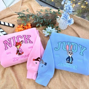 May include: Pink and light blue sweatshirts featuring characters from Zootopia. The pink sweatshirt has "NICK" embroidered on it, with a fox graphic. The light blue sweatshirt has "JUDY" embroidered on it, with a bunny graphic. Both have "ZOOTOPIA" embroidered.