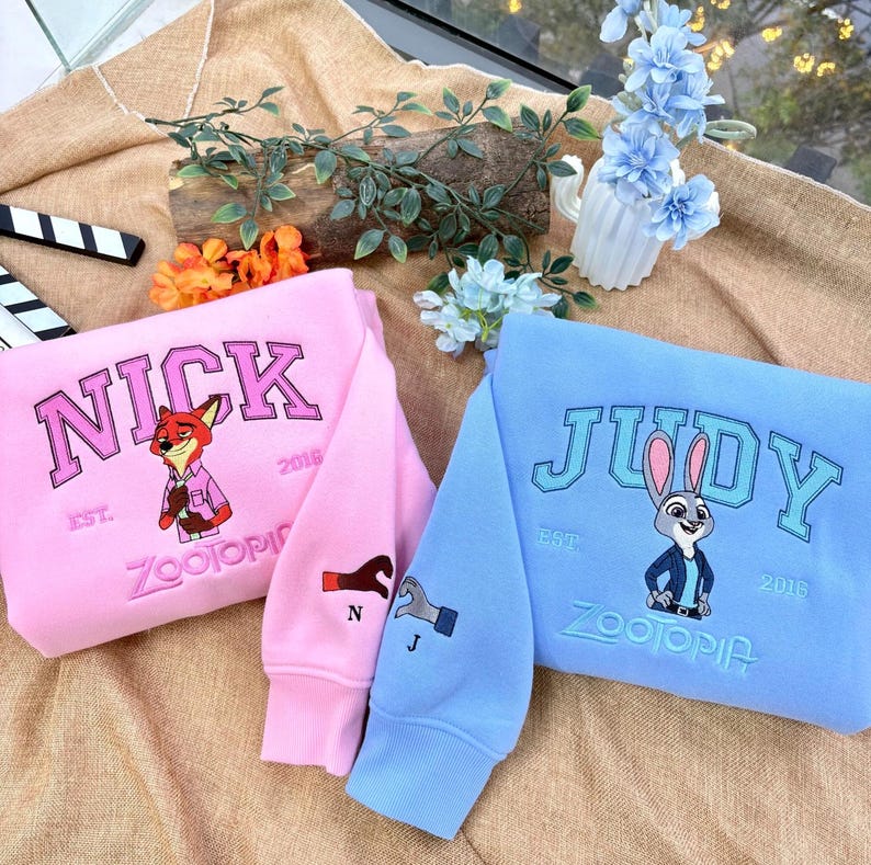 May include: Pink and light blue sweatshirts featuring characters from Zootopia. The pink sweatshirt has "NICK" embroidered on it, with a fox graphic. The light blue sweatshirt has "JUDY" embroidered on it, with a bunny graphic. Both have "ZOOTOPIA" embroidered.